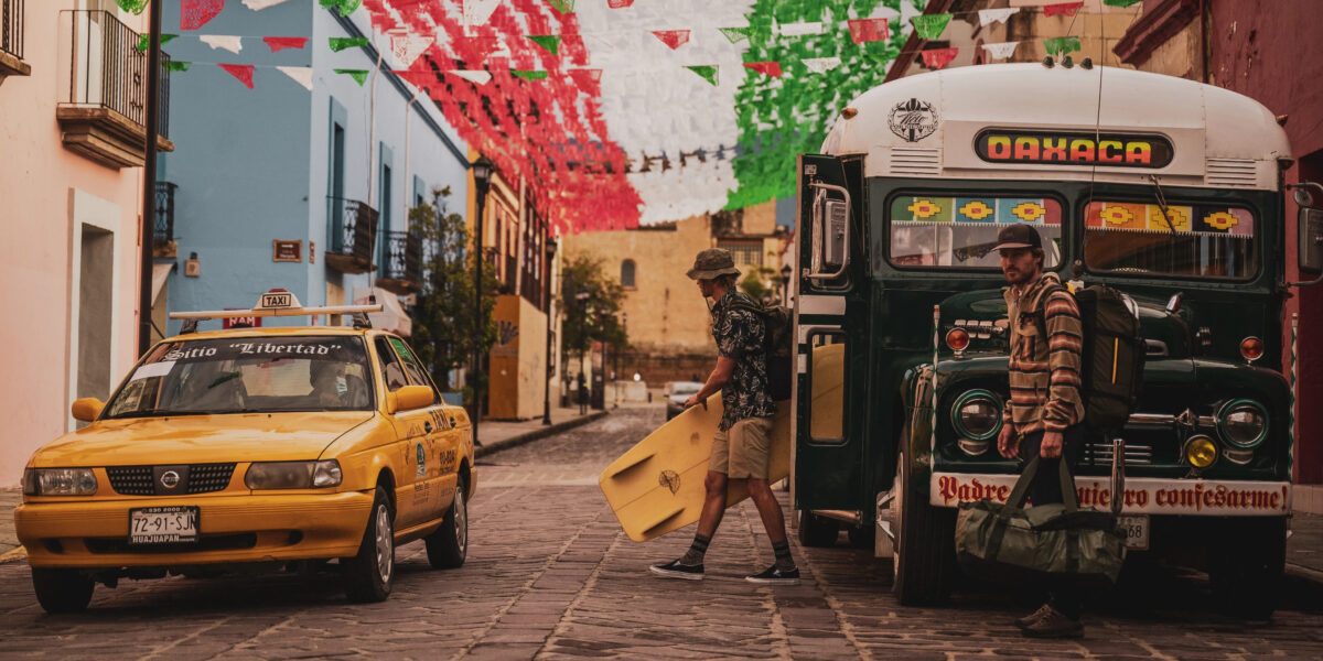 Roark in Oaxaca Mexico, two surfer dudes dressed in Roark clothing, carrying a surfboard in front of a bus and taxi